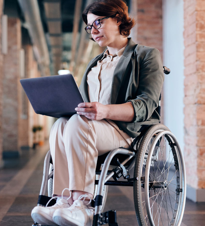 A person wearing spectacles is looking at their computer on their lap. They are sitting in a wheelchair and the background is unclear but looks corporate, like an office.