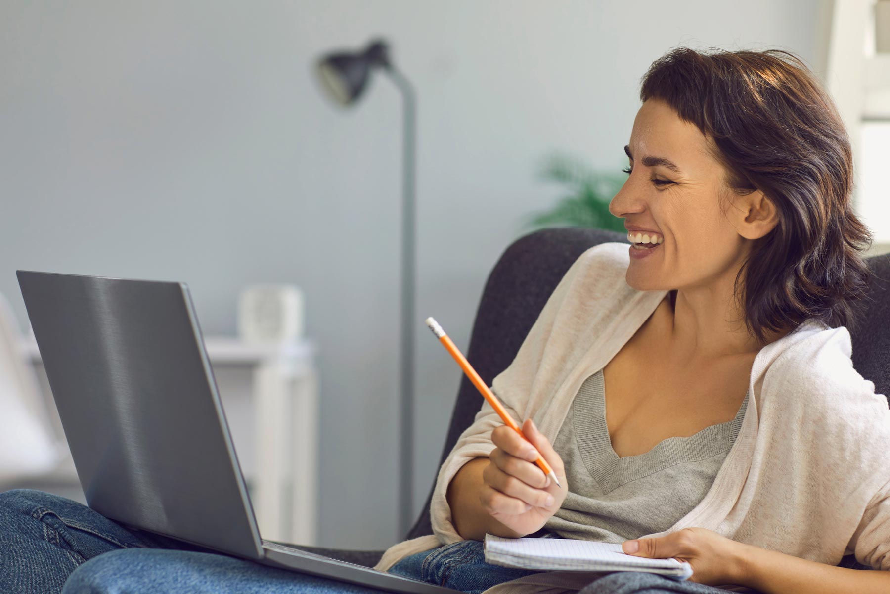 Person is sitting on a grey sofa in their living room holding a notepad and looking at a computer screen on their lap.