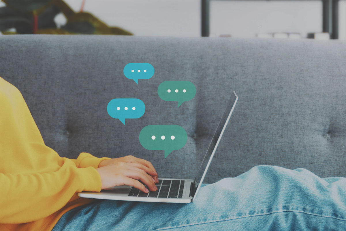 A person is sitting on a grey sofa in a yellow jumper and blue jeans. We cannot see their face. They have a laptop on their legs and some green speech bubbles have been added to the image above the laptop to demonstrate that they are messaging somebody.