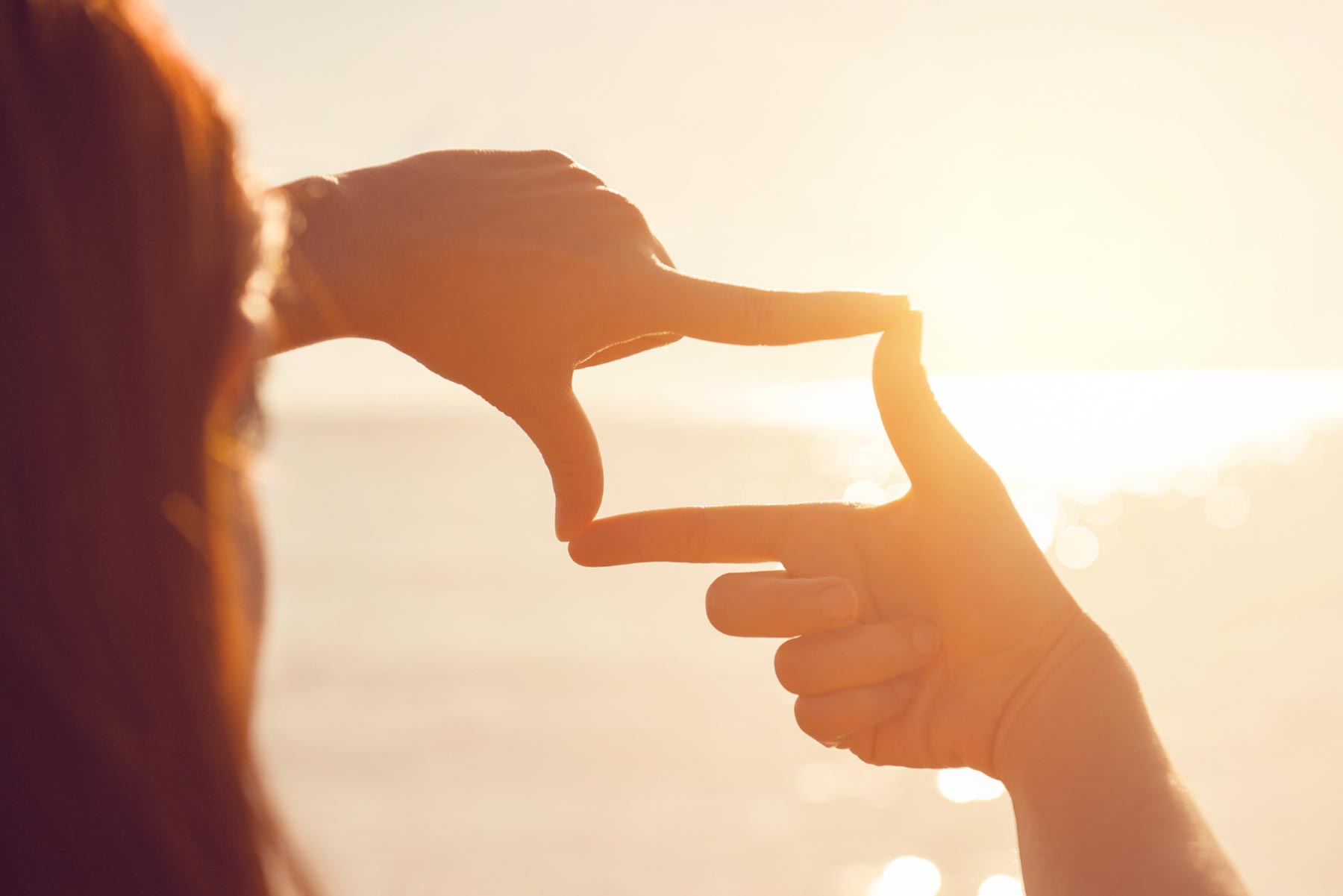 Photo shows a small part of the side of a person's head, with their back to the camera. The photo is mainly focused on the hands of this individual, which they are using to frame the sunset.