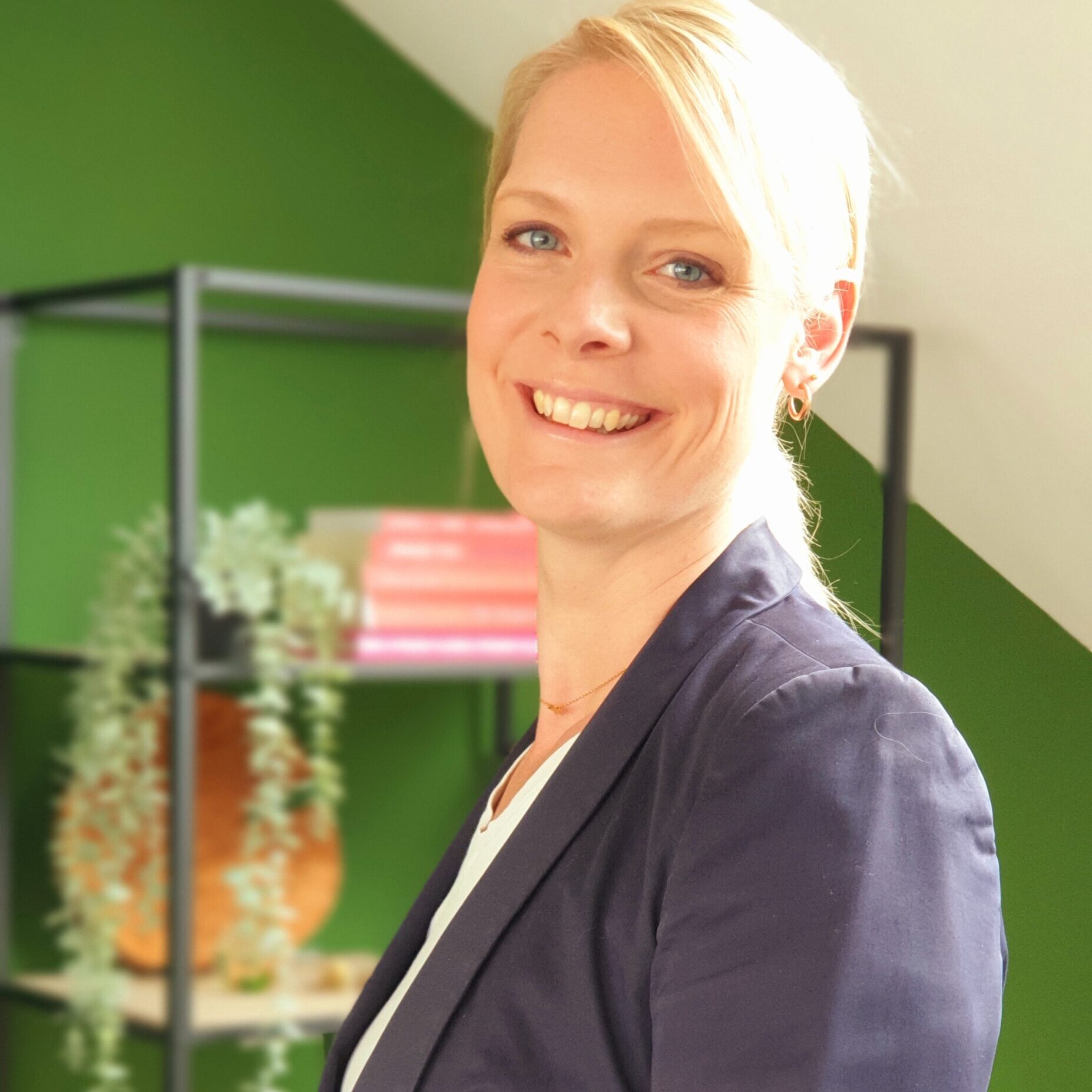 20210623_102138 A woman in her forties with light skin and blond hair. The sun is shining on her hair. She is looking towards the camera and smiling. She is wearing a blue formal jacket. The background is a green wall with a bookcase and some red books.
