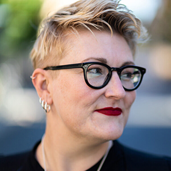 A white nonbinary person (Alex Haagarrd) with short blond hair looking off to the side and smiling slightly. They are wearing eyeglasses with round, black plastic frames, dark red lipstick and four silver hoop earrings in progressively smaller sizes.