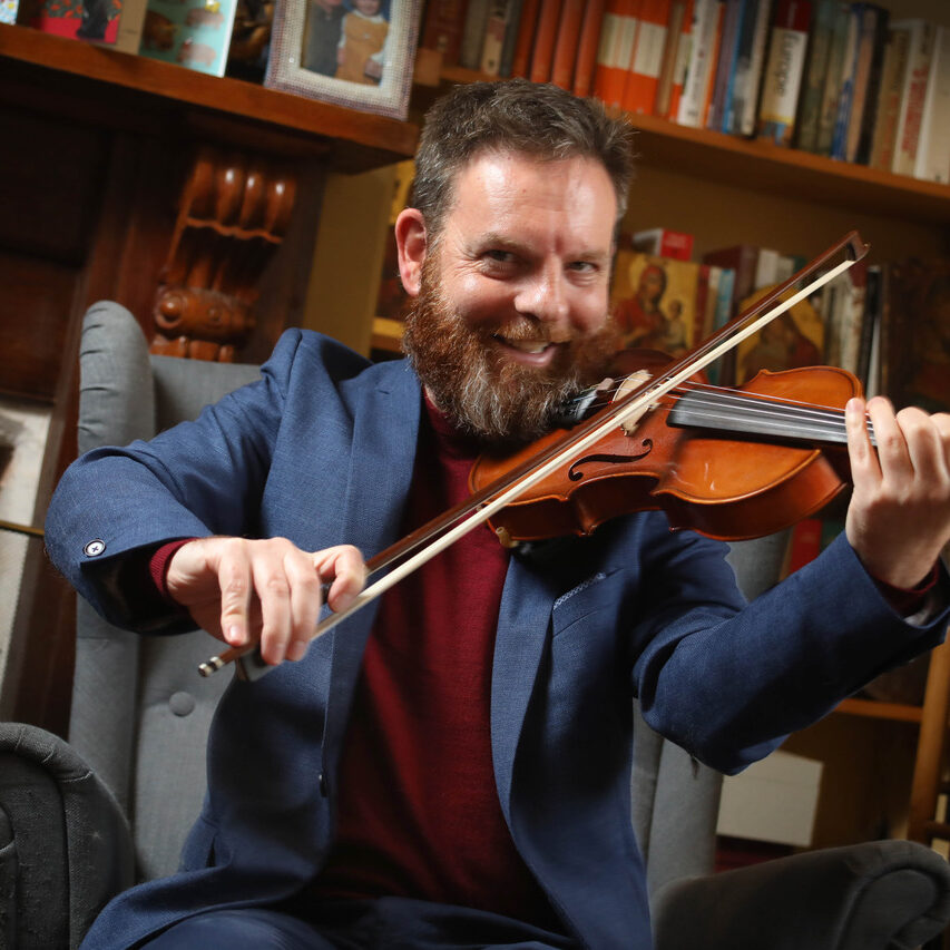A bearded male-presenting Caucasian person playing the violin. He is seated in an armchair. In the background is a bookshelf full of books, and in the foreground is an iPad sitting on a music stand, with an Augmentative and Accessibility app on the screen.