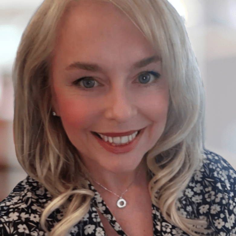 A middle-aged white woman with long, blonde hair is smiling warmly at the camera. She’s wearing a black blouse with a white floral pattern and a silver necklace. This is a professional headshot reflecting the confidence and approachability she brings to the health advocacy space.