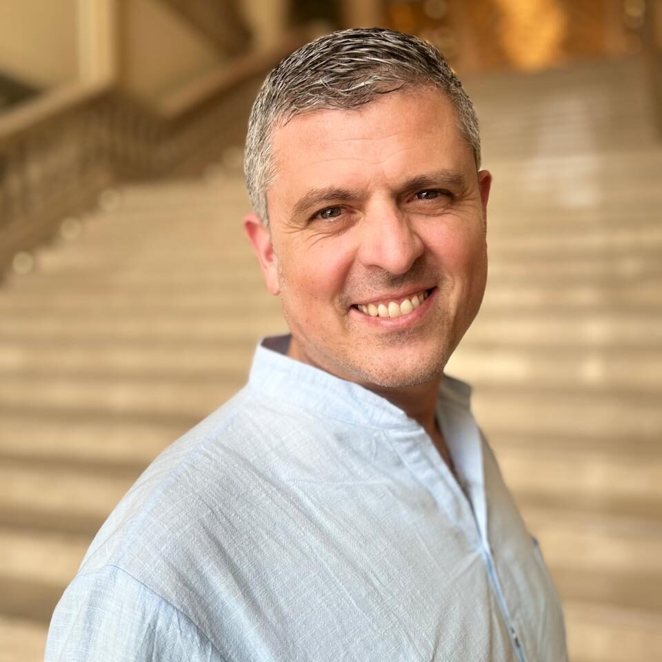 A white man in his forties with short, salt-and-pepper hair and no facial hair. He is smiling warmly and wearing a light blue, collarless shirt. His body is turned slightly to the side, and the background shows a large, elegant staircase in a well-lit indoor setting.