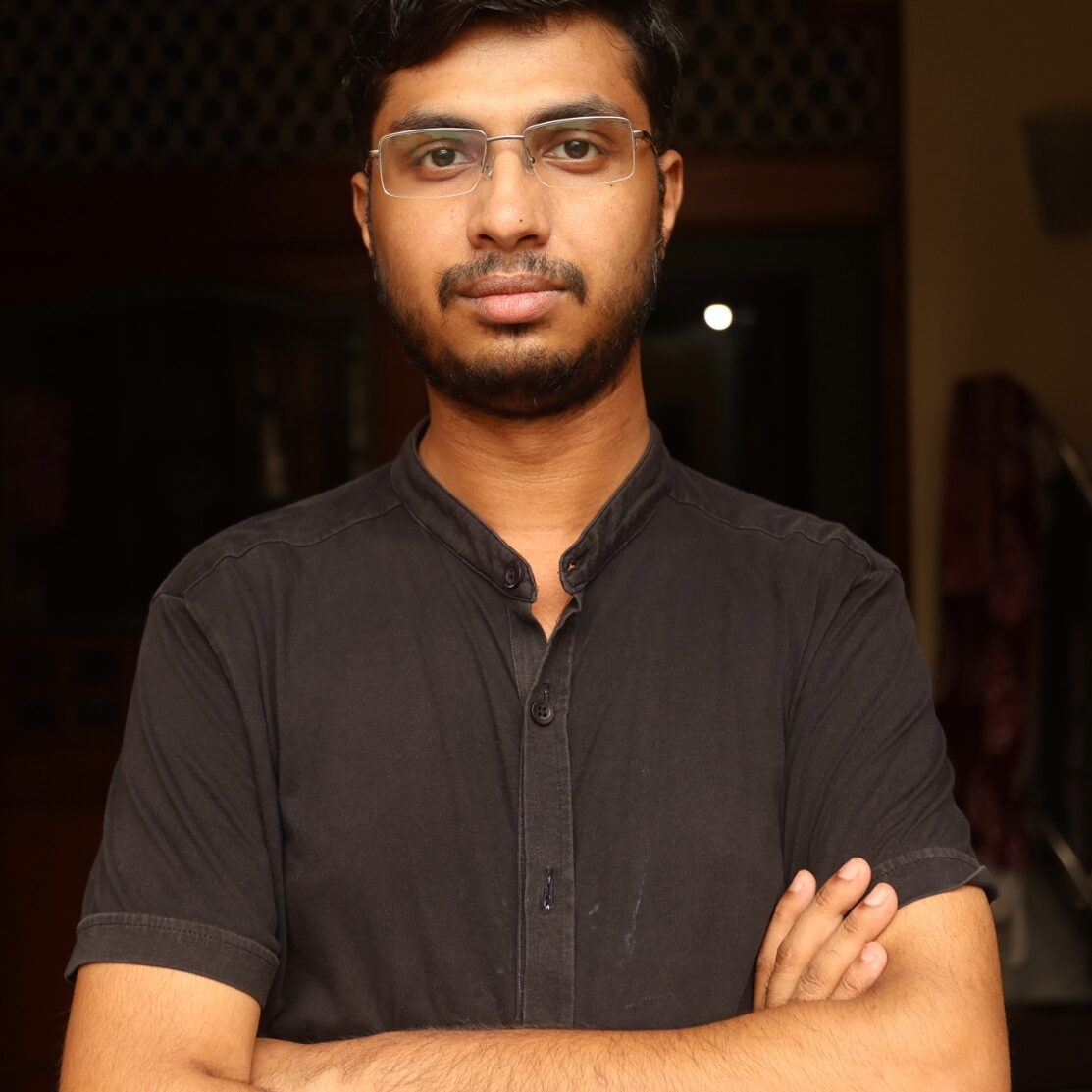 A confident-looking man with medium-brown skin, short black hair, a trimmed beard, and rectangular glasses stands indoors with his arms crossed. He is wearing a black mandarin-collar short-sleeved shirt and gazes directly at the camera with a neutral expression. The background appears to be a warmly lit room with wooden and patterned decor elements.