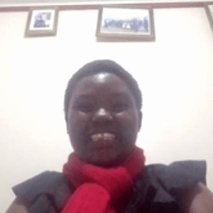 Screenshot 2025-08-01 164014 A person smiles at the camera, wearing a red scarf and dark top, with framed pictures and a calendar on the wall behind them.