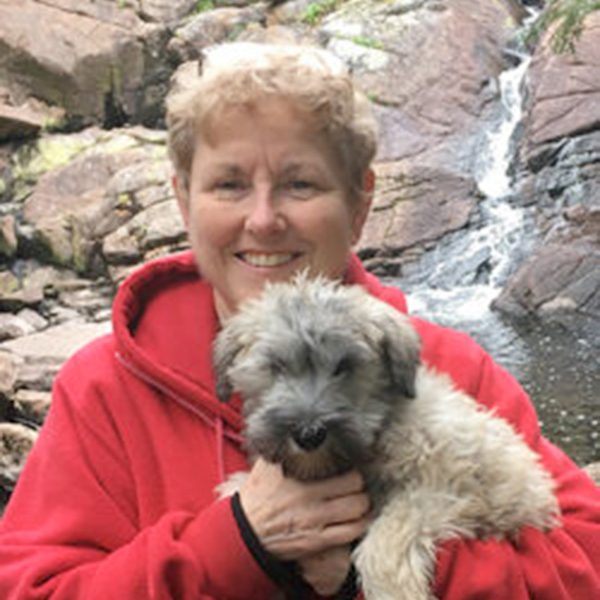 This is a photo of Trudy Flynn, smiling and holding a small dog. There is a waterfall and rocks in the background.